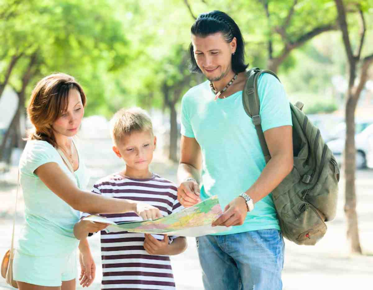 A family stands in a sunny park, studying a map together, surrounded by green trees and parked cars in the background.