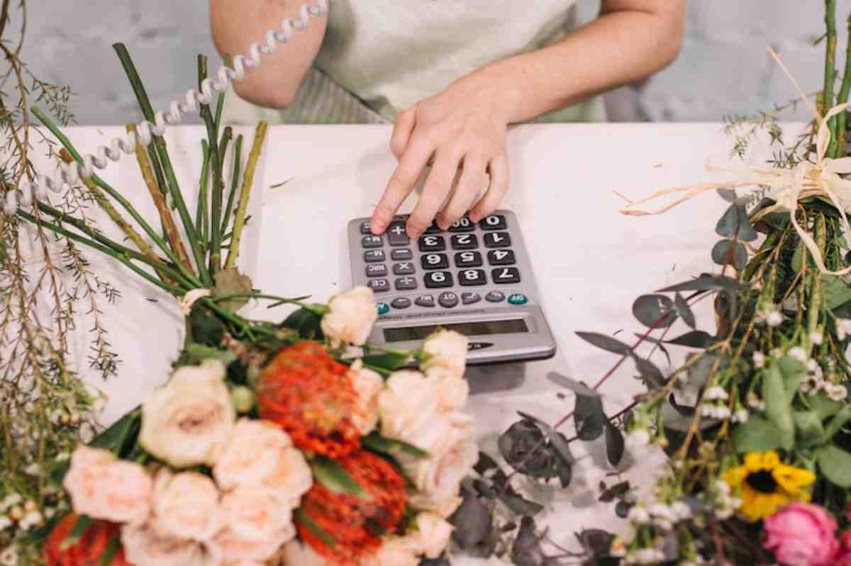 A person calculates costs using a calculator, surrounded by vibrant flower arrangements on a table.