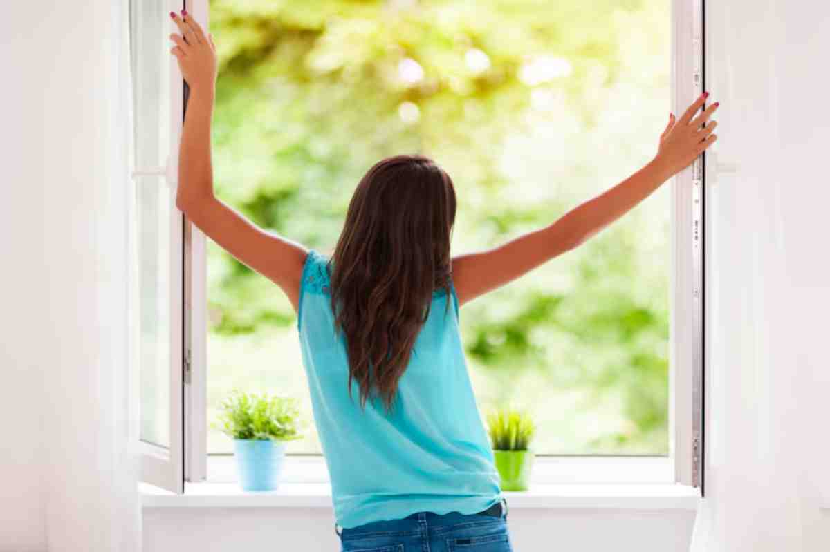A girl in a blue tank top stands at an open window, stretching her arms wide, with green plants on the windowsill and sunlight streaming in.