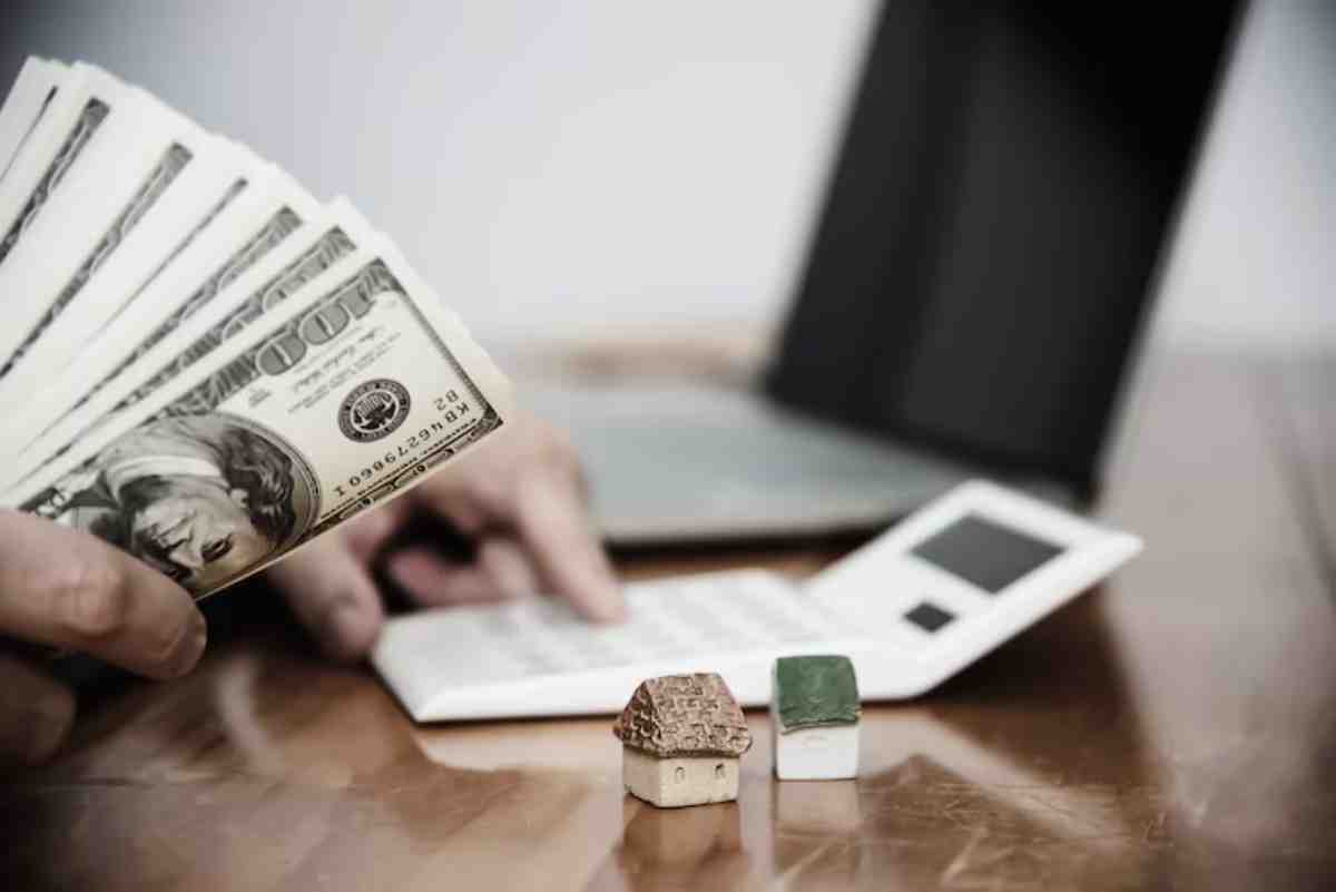 A person holds a fan of $100 bills next to a calculator and two toy houses, symbolizing real estate finance and investment.