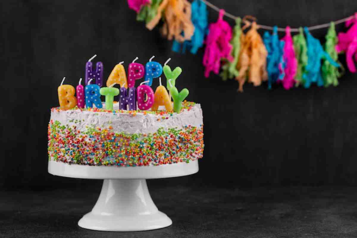 A colorful birthday cake with Happy Birthday candles, surrounded by sprinkles, displayed on a white pedestal against a dark background.