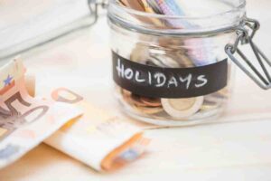 A glass jar labeled Holidays filled with coins and cash notes sits alongside a €50 bill on a light wooden surface.