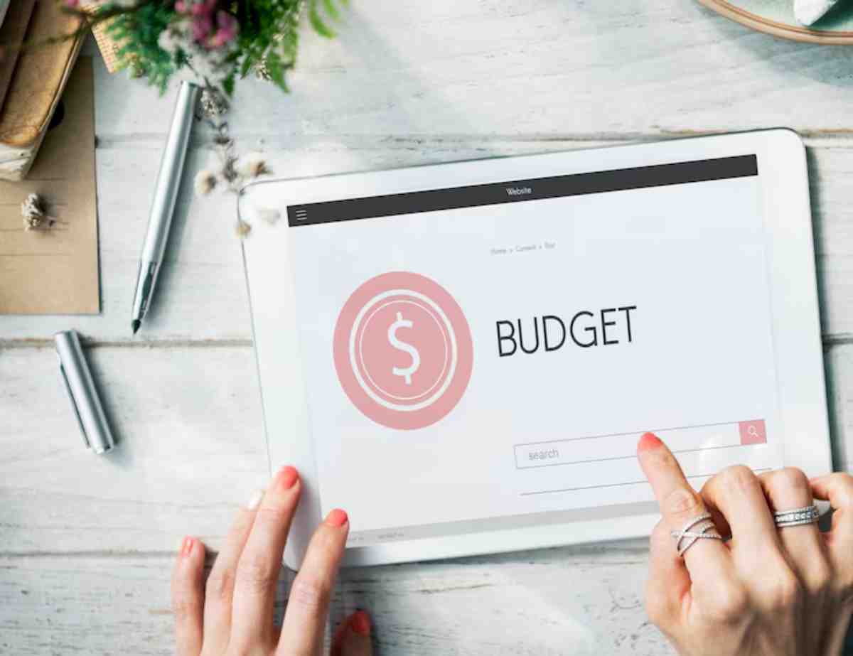 A hand with pink nails navigates a tablet displaying a budgeting app, featuring a dollar symbol and a search bar.