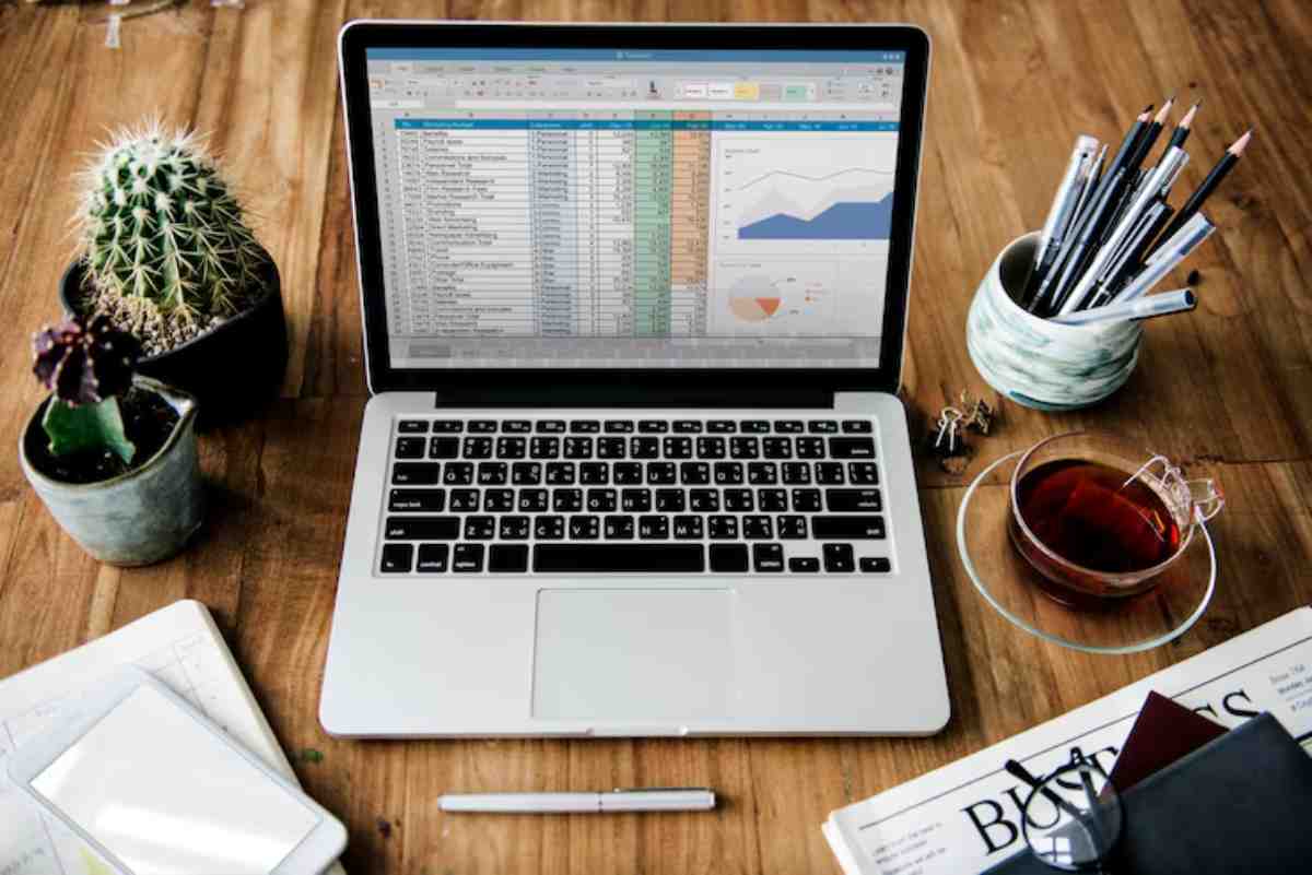 A laptop displaying a spreadsheet and graphs, surrounded by plants, a cup of tea, pens, and a notepad on a wooden desk.