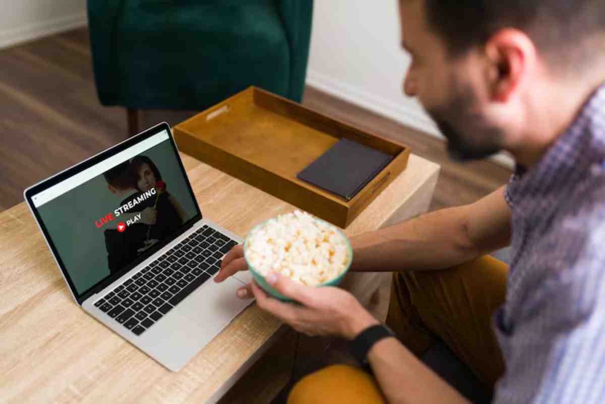 A person holds a bowl of popcorn while using a laptop displaying a streaming service, preparing to watch content.