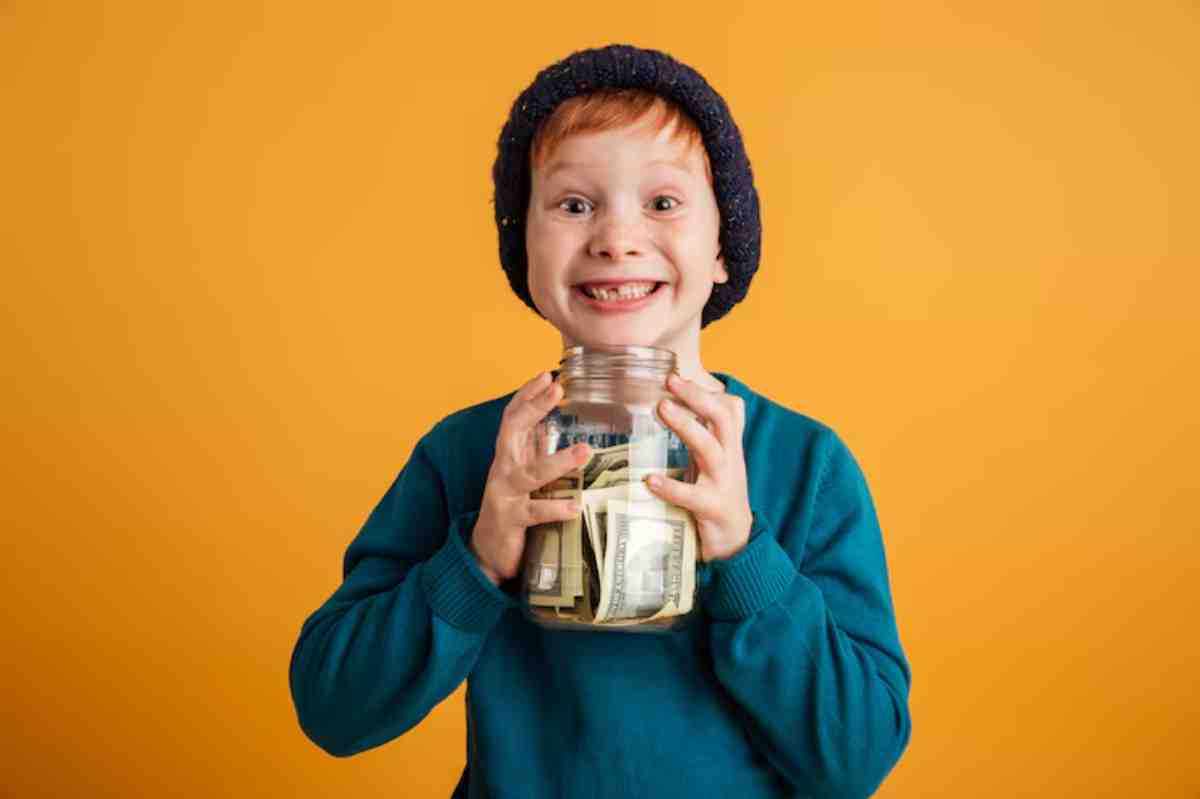 A child holds a jar filled with cash, wearing a green sweater and a knitted hat, against a vibrant orange background.