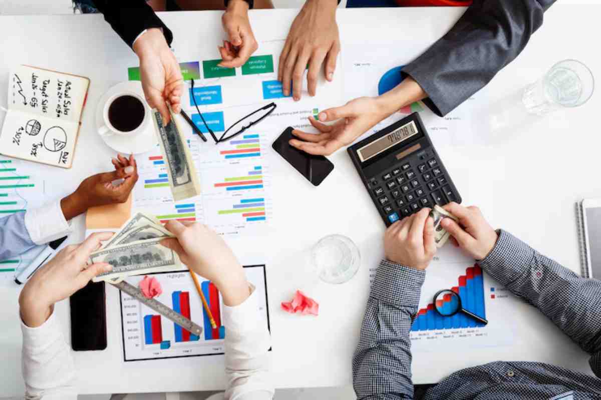 Hands exchanging cash over a table covered with financial charts, a calculator, and documents, indicating a business discussion.