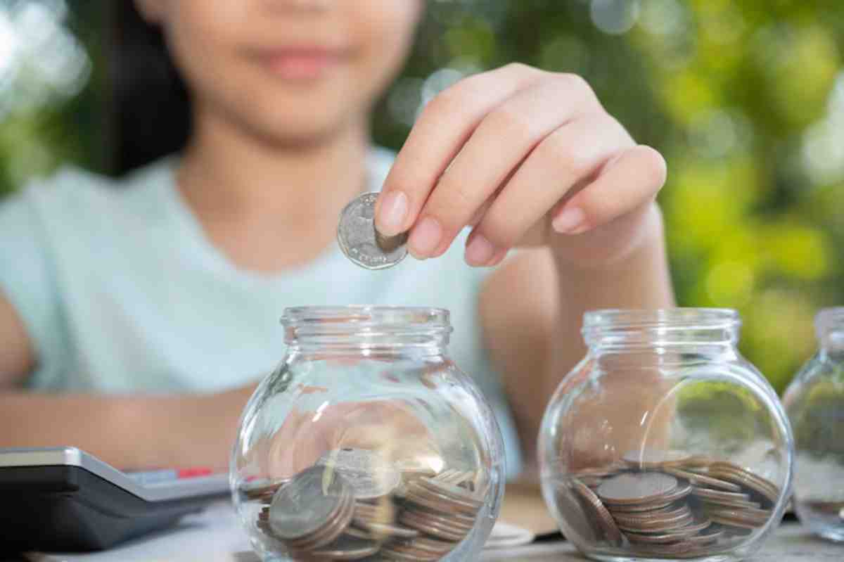 A girl inserts a coin into a glass jar filled with coins, surrounded by greenery and a calculator on the table.