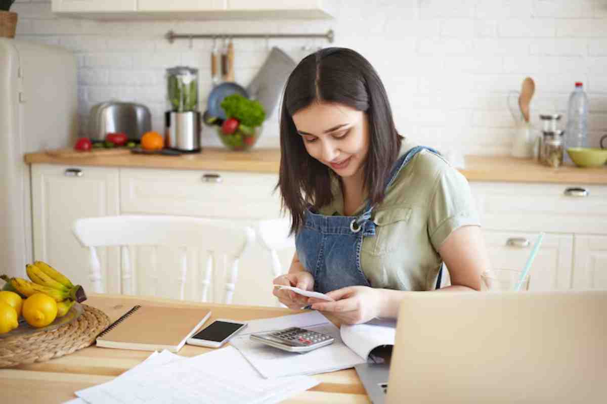 A young woman in denim overalls sits at a kitchen table with a calculator, papers, a smartphone, and fresh fruit nearby.