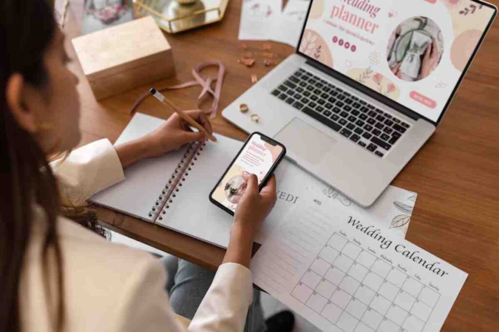 A person writes notes in a notebook while holding a smartphone displaying wedding planning details, with a laptop and calendar visible.