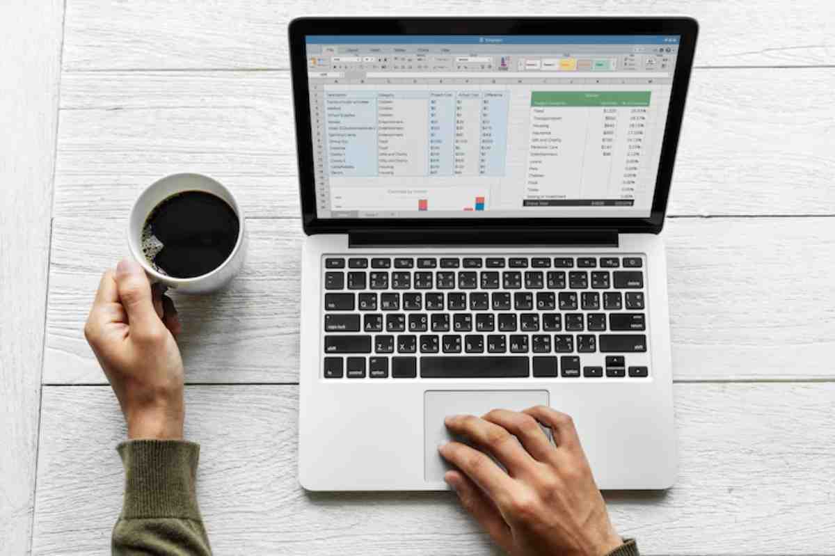 A person sips coffee while working on a laptop with an open Excel spreadsheet, placed on a wooden table.
