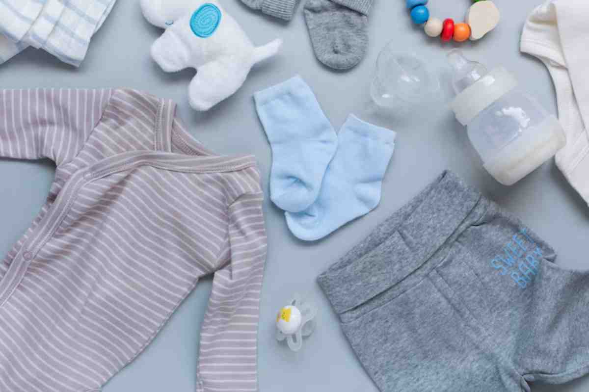 A flat lay of baby items: a striped onesie, socks, plush toy, pacifier, bottle, blanket, and colorful teething ring on a light background.