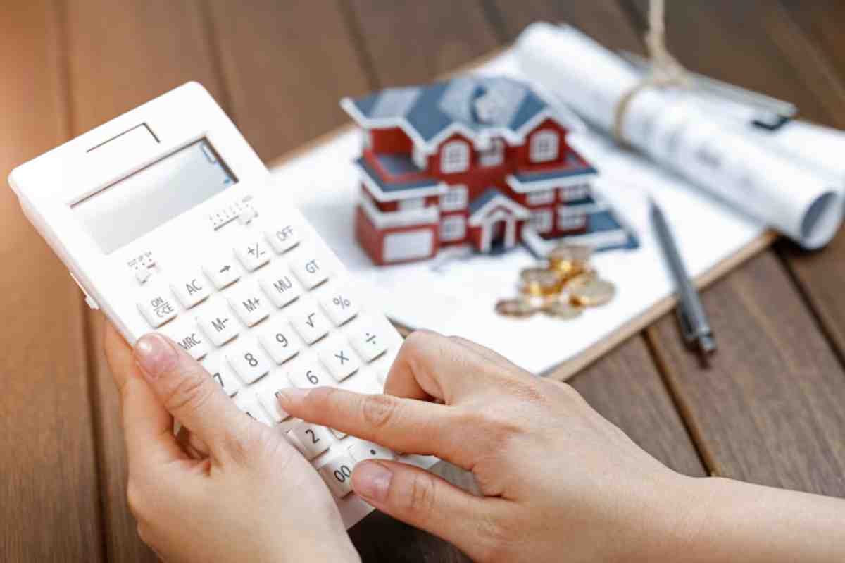 A person calculates finances with a calculator, alongside a model house, blueprints, and coins on a wooden table.