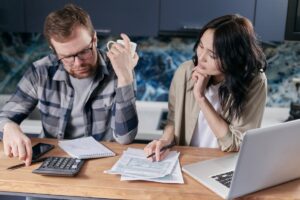 A man and a woman sit at a table covered with paperwork, a laptop, and a calculator, engaged in a serious discussion.