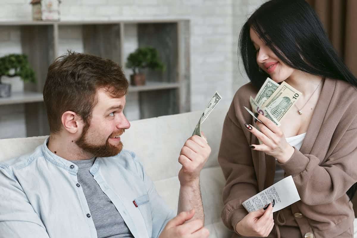A man and woman sit on a sofa discussing money, with the woman holding cash and a receipt, appearing engaged in a conversation.