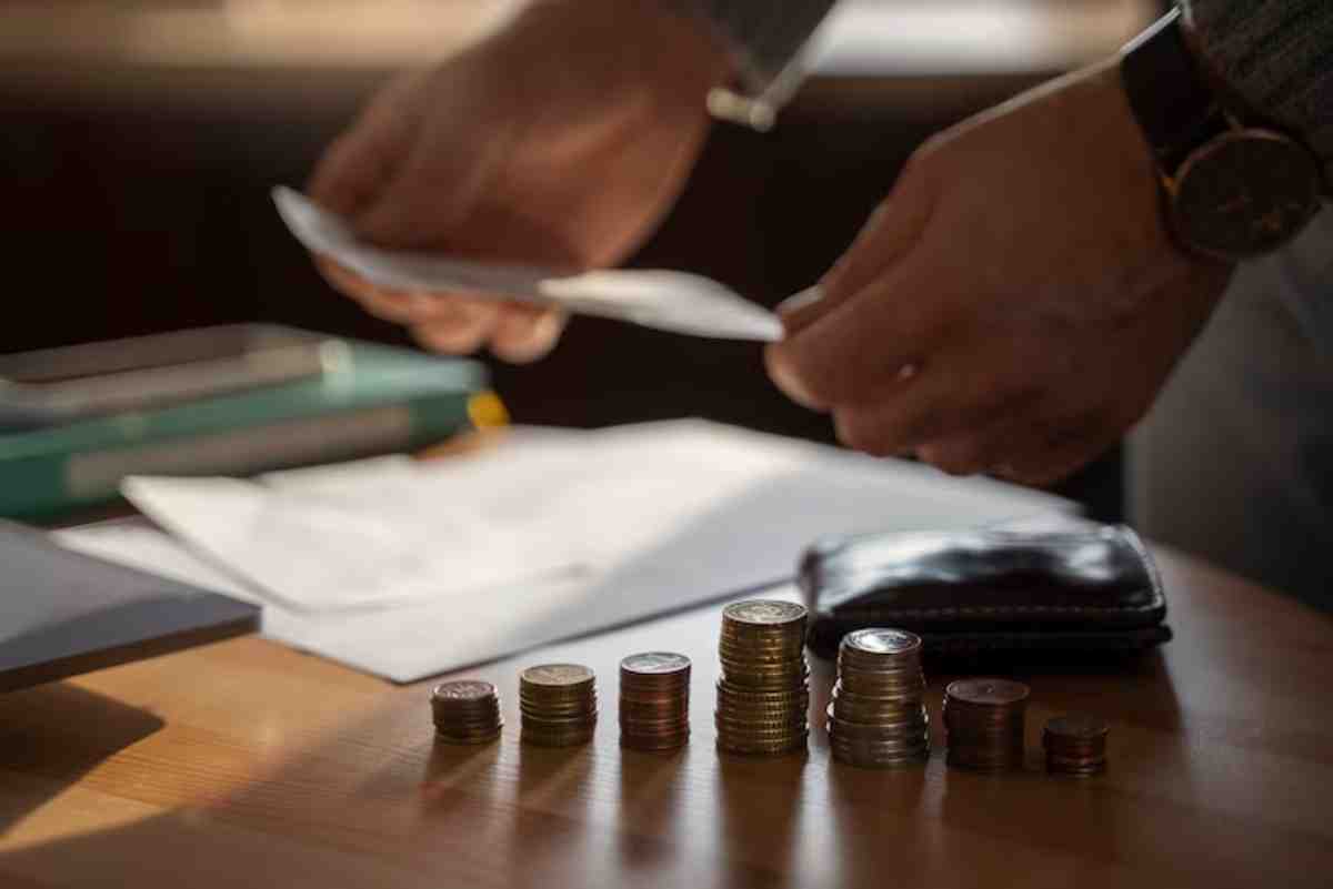 A person organizes coins into stacks on a wooden table, preparing to handle papers and money. A wallet and phone are nearby.