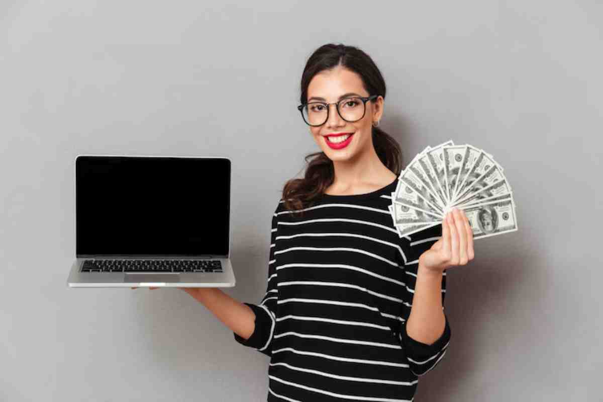 A person in a striped shirt holds a laptop in one hand and a fan of cash in the other, against a grey background.