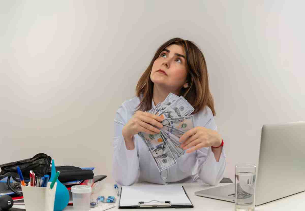 A person holds bundles of cash while sitting at a cluttered desk with a laptop, stationery, and a glass of water.