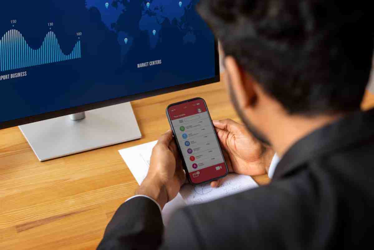 A businessman reviews data on his smartphone while analyzing market trends displayed on a monitor in an office setting.
