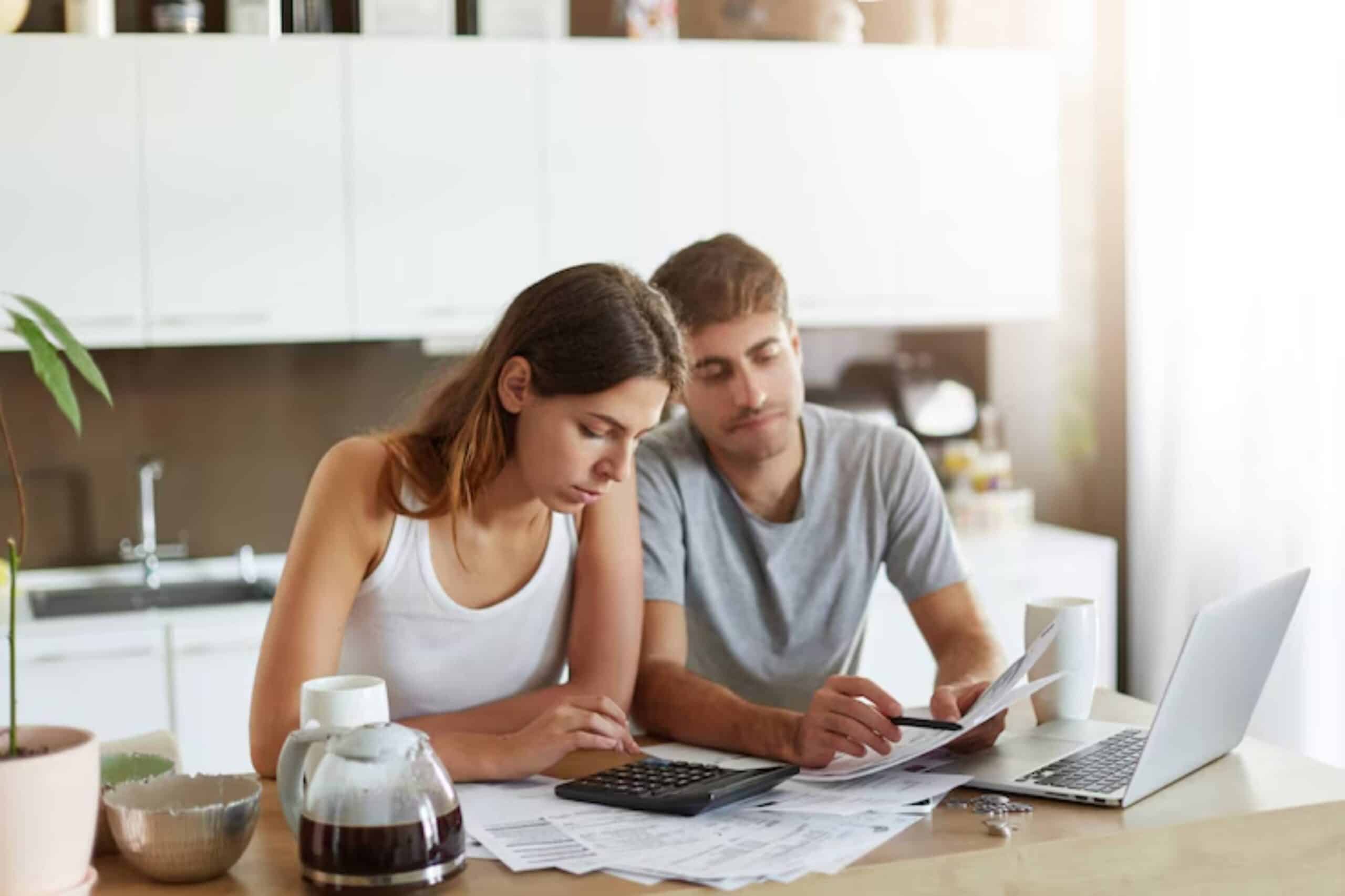 A couple sits at a kitchen table, reviewing documents and using a calculator, with coffee and a laptop nearby.